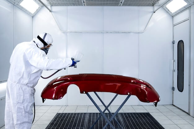 Painter applying new layer of fresh metallic paint on the automobile bumper in painting chamber.