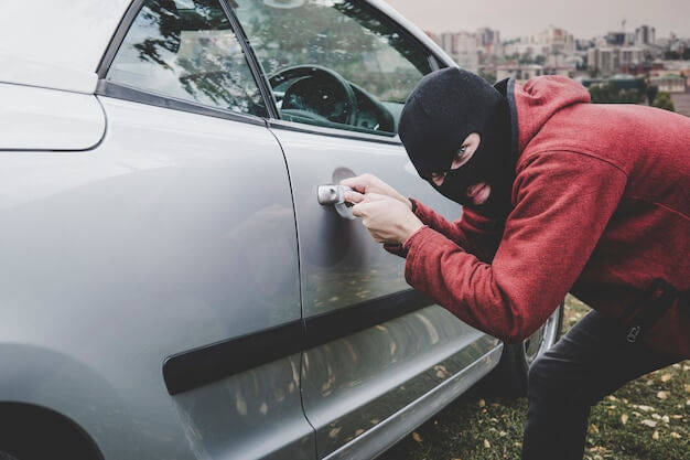 Photo thief looking inside a car window ready to steal something. criminal in black balaclava and hoodie opens somebody's vehicle with skeleton key. car breaking by unknown male with hidden face.